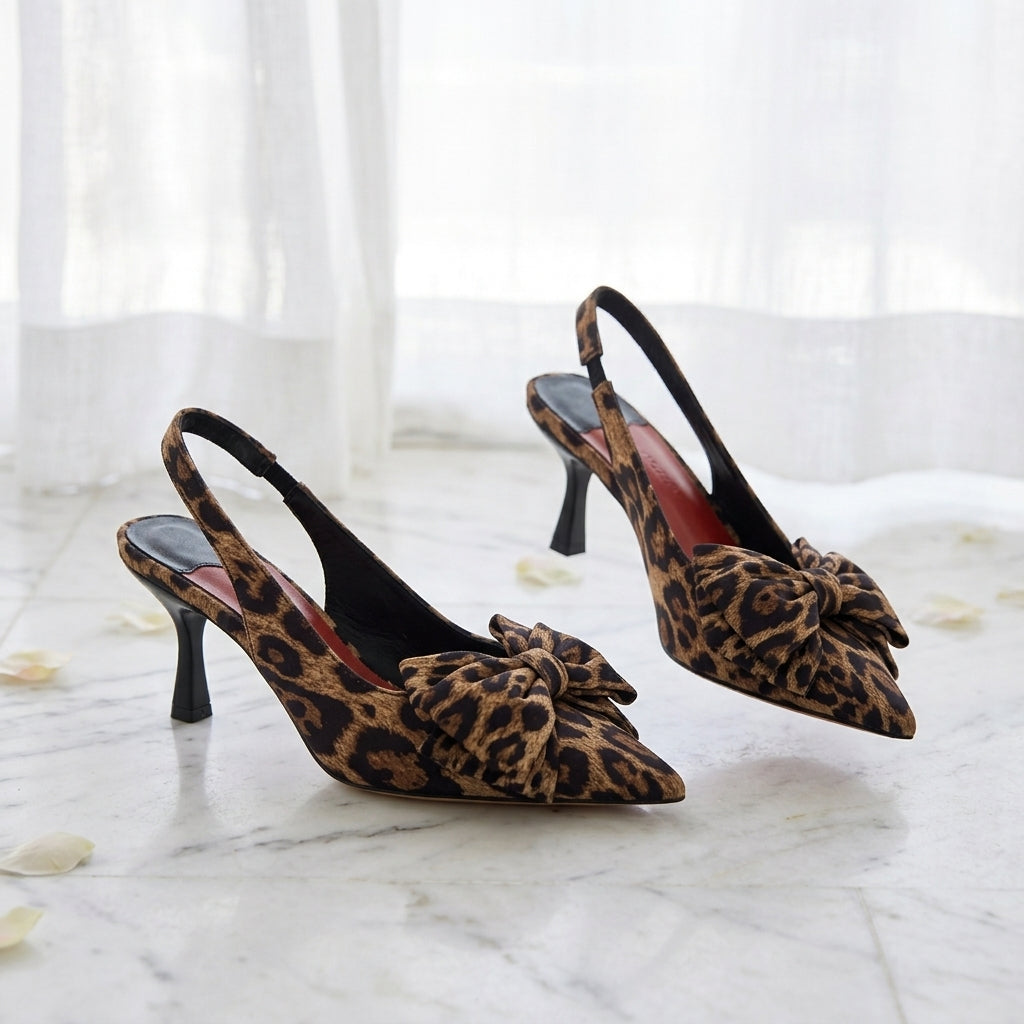 A pair of pointed-toe leopard print slingback heels featuring a decorative bow on the front, displayed on a white marble surface.
