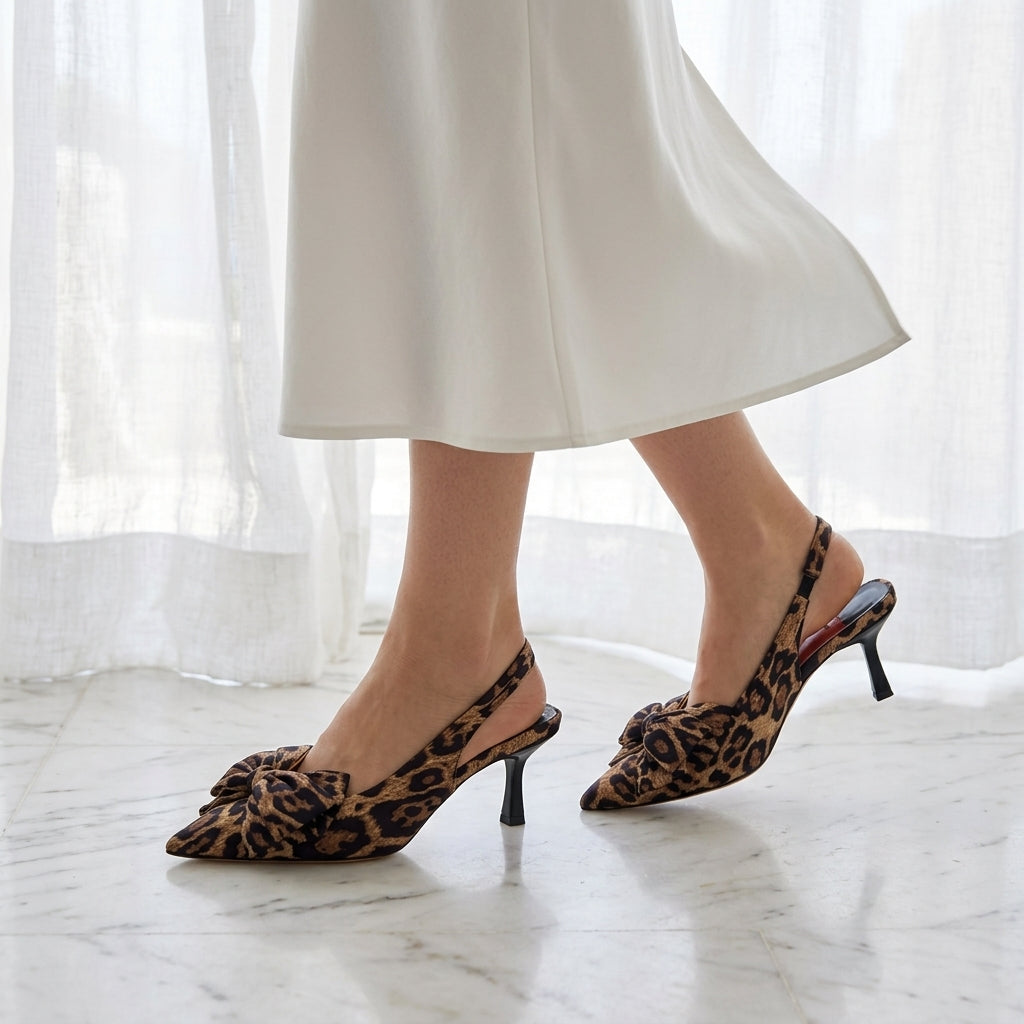 A pair of leopard-print pointed-toe slingback heels featuring a decorative bow on the front, worn with a white skirt on a marble floor.