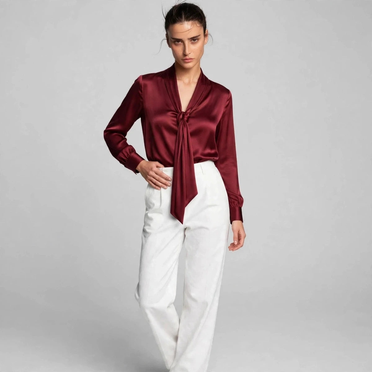 Model wearing burgundy satin tie-neck blouse with white trousers in a fashion editorial