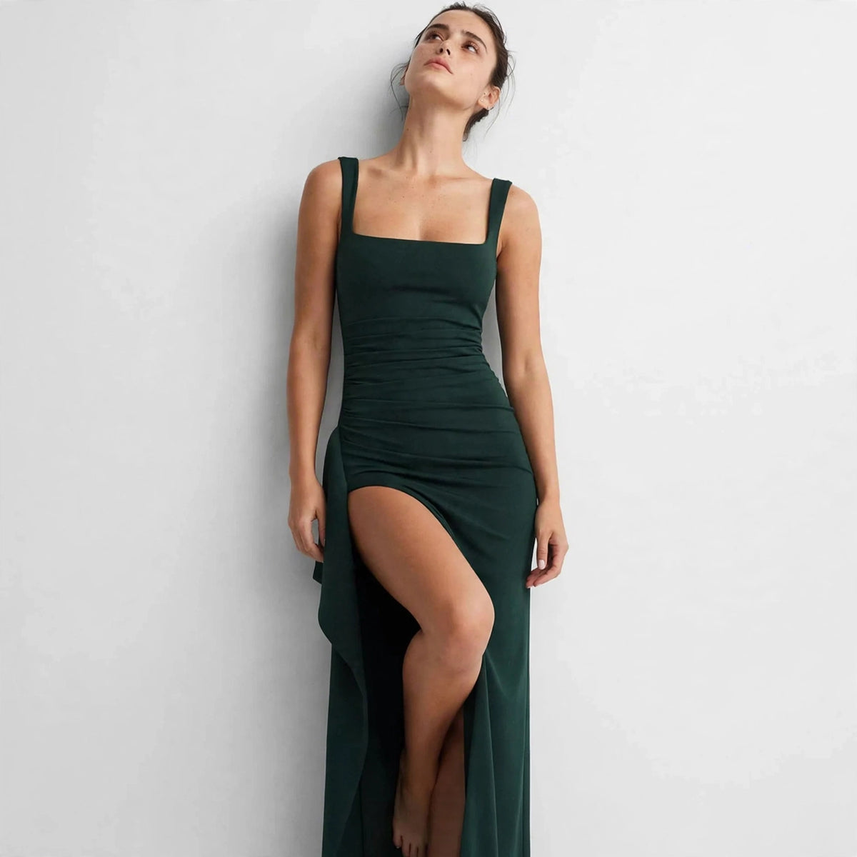 Model leaning against a wall in a dark green ruched high-slit maxi dress, front view