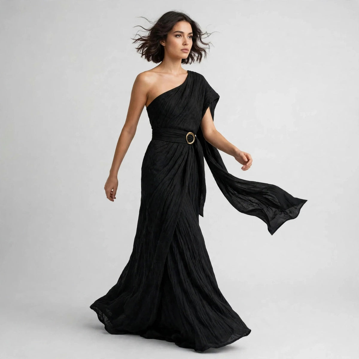Model walking in a black draped one-shoulder maxi dress with a belted ring buckle, skirt flowing.