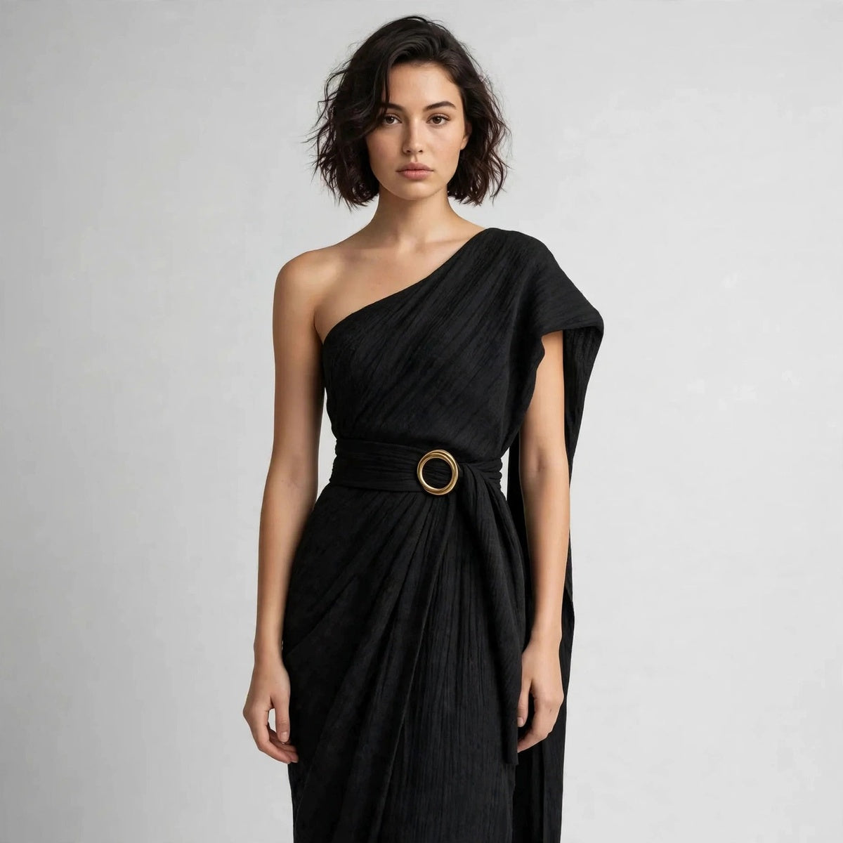 Close-up of a model in a black draped one-shoulder dress with a wide belt and round ring buckle at the waist.