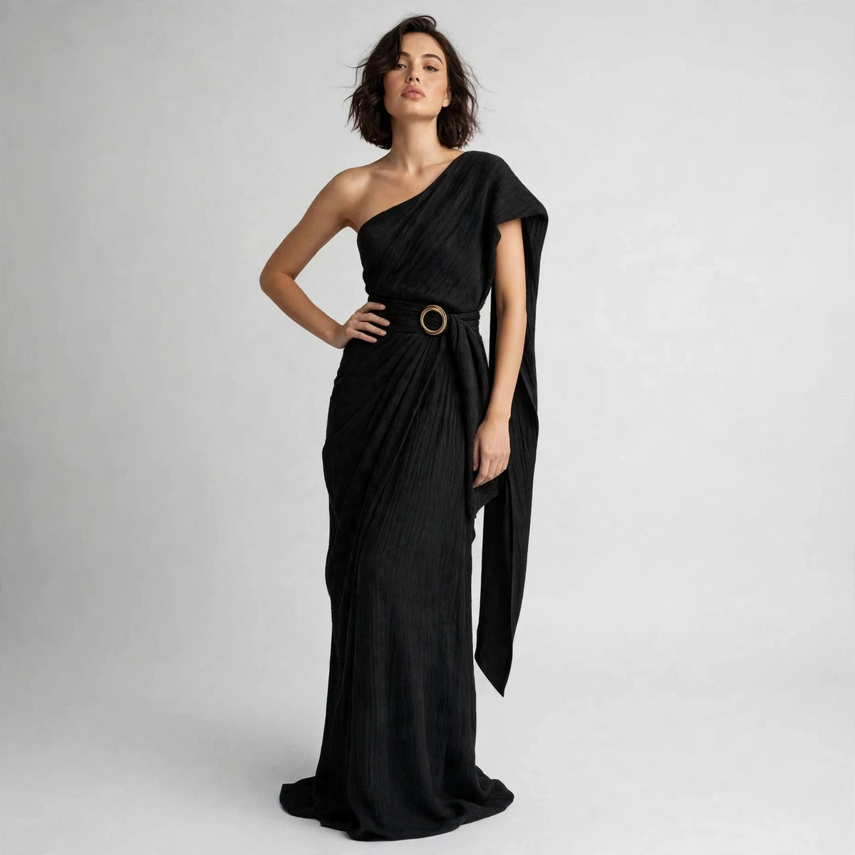 Model standing with hand on hip in a black draped one-shoulder maxi dress with a belted ring buckle.