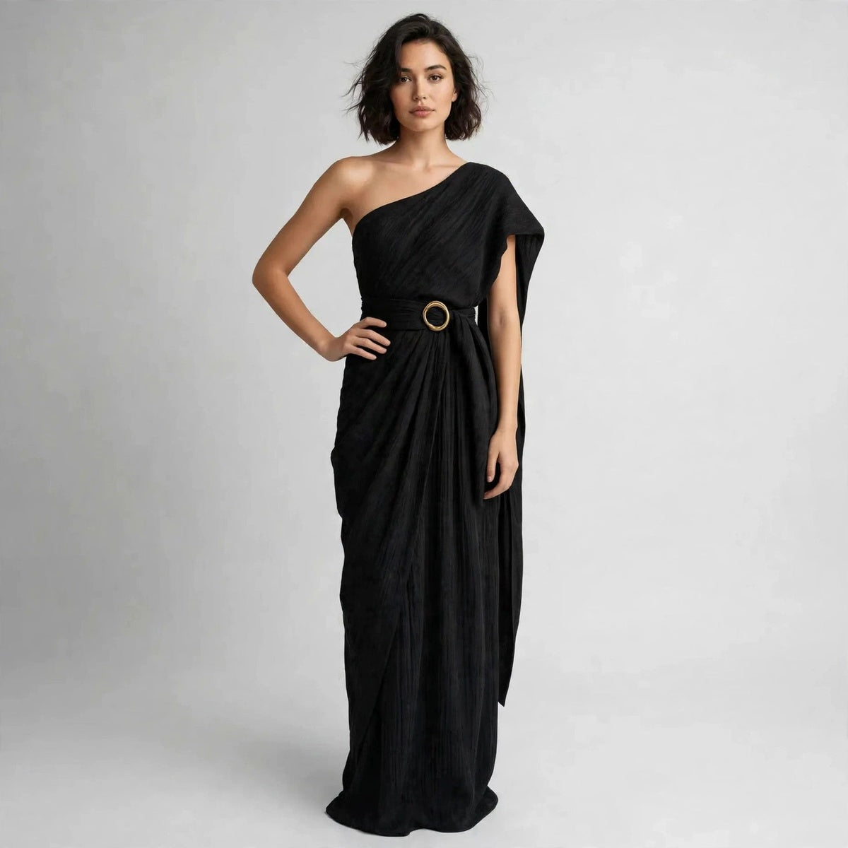 Full-length view of a model with hand on hip in a black draped one-shoulder maxi dress with a belted ring buckle.