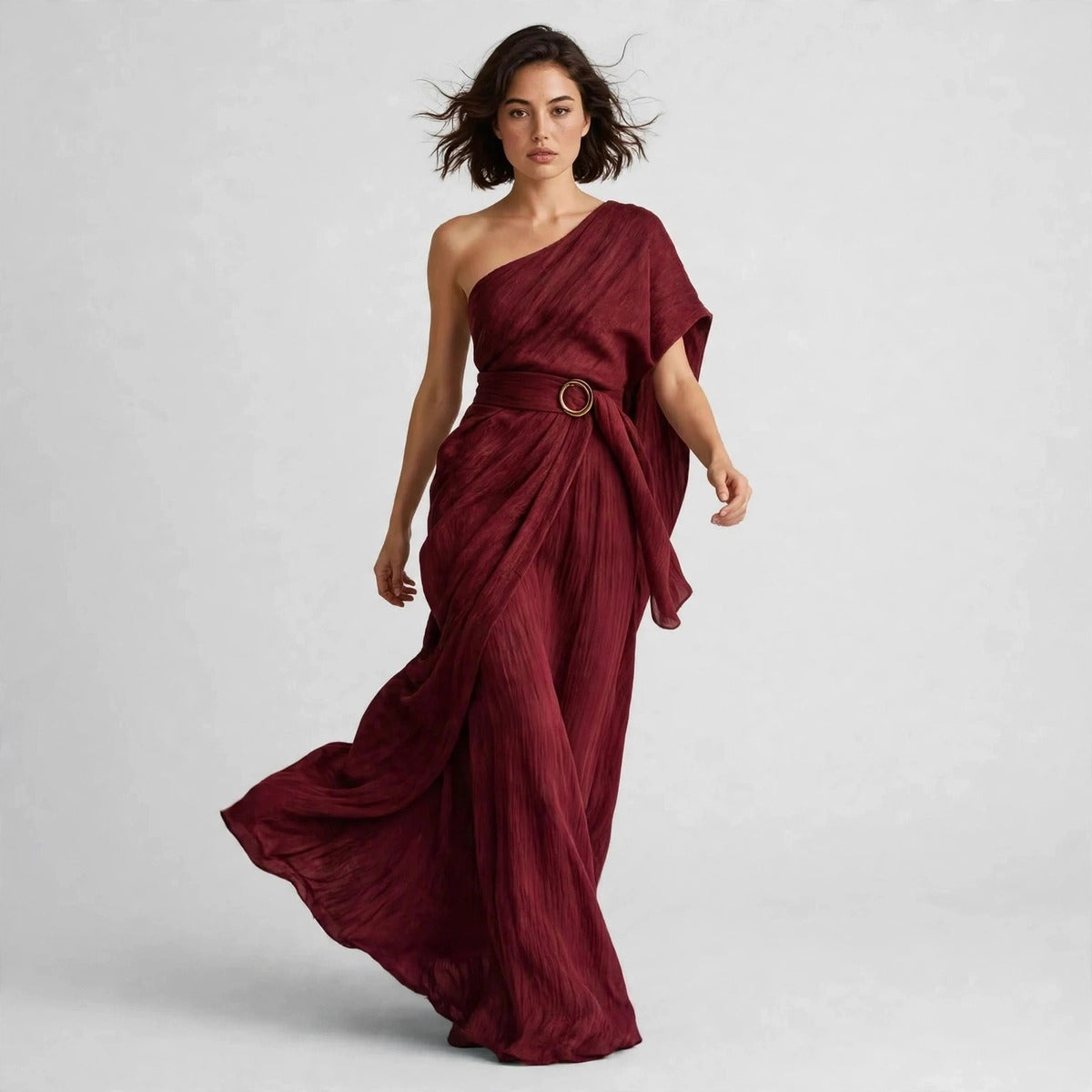 Model walking in a burgundy draped one-shoulder maxi dress with a belted ring buckle, skirt flowing.