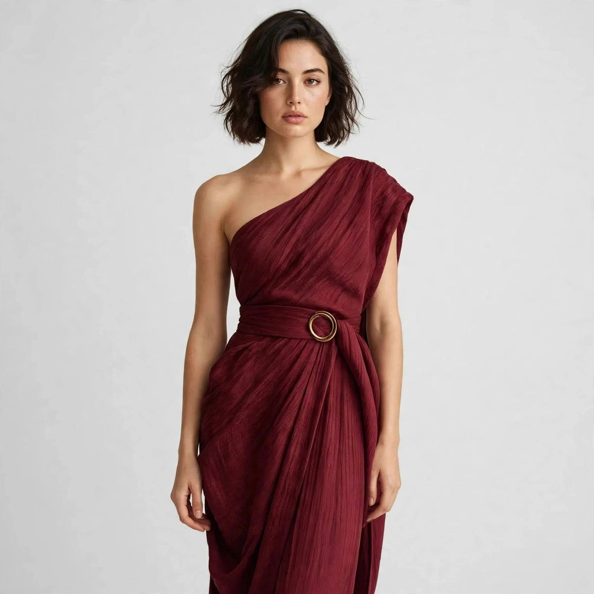 Close-up of a model in a burgundy draped one-shoulder dress with a wide belt and round ring buckle at the waist.