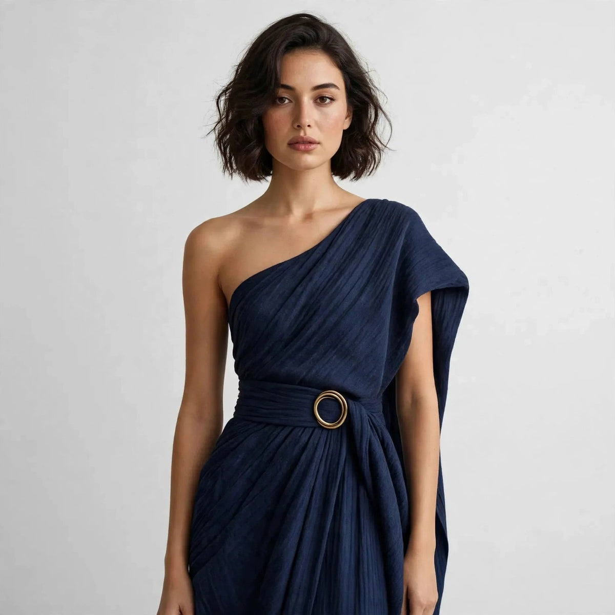 Close-up of a model in a navy blue draped one-shoulder dress with a wide belt and round ring buckle at the waist.