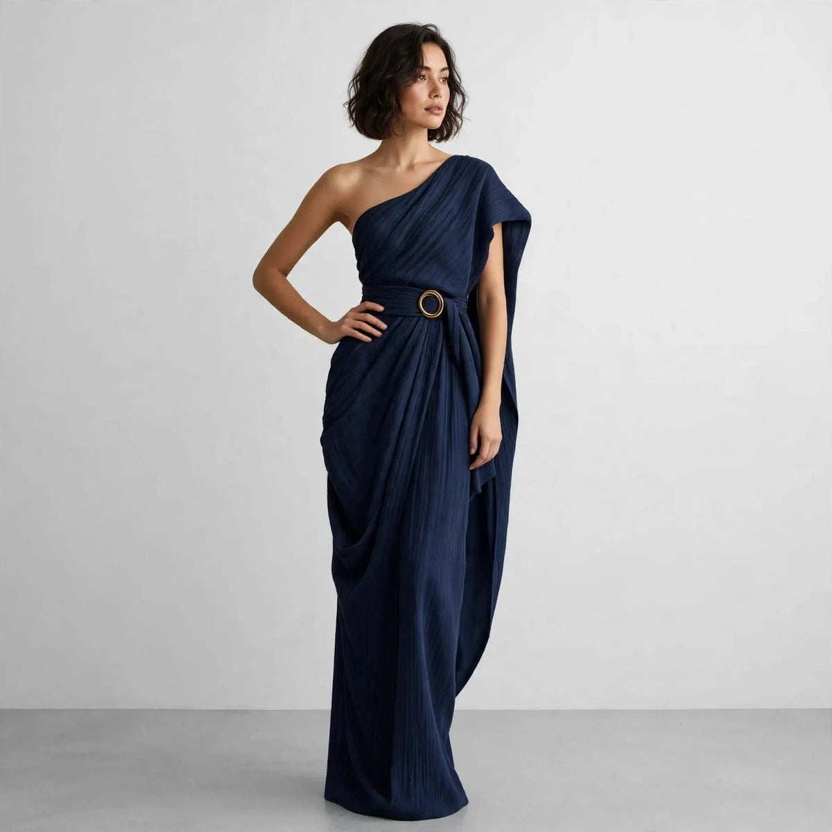 Full-length view of a model with hand on hip in a navy blue draped one-shoulder maxi dress with a belted ring buckle.