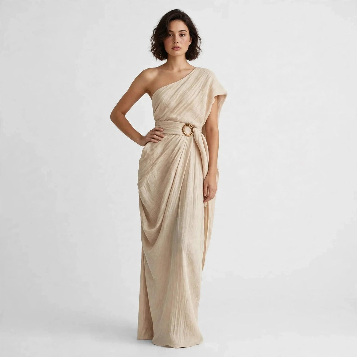 Model standing with hand on hip in a tan draped one-shoulder maxi dress with a belted ring buckle.