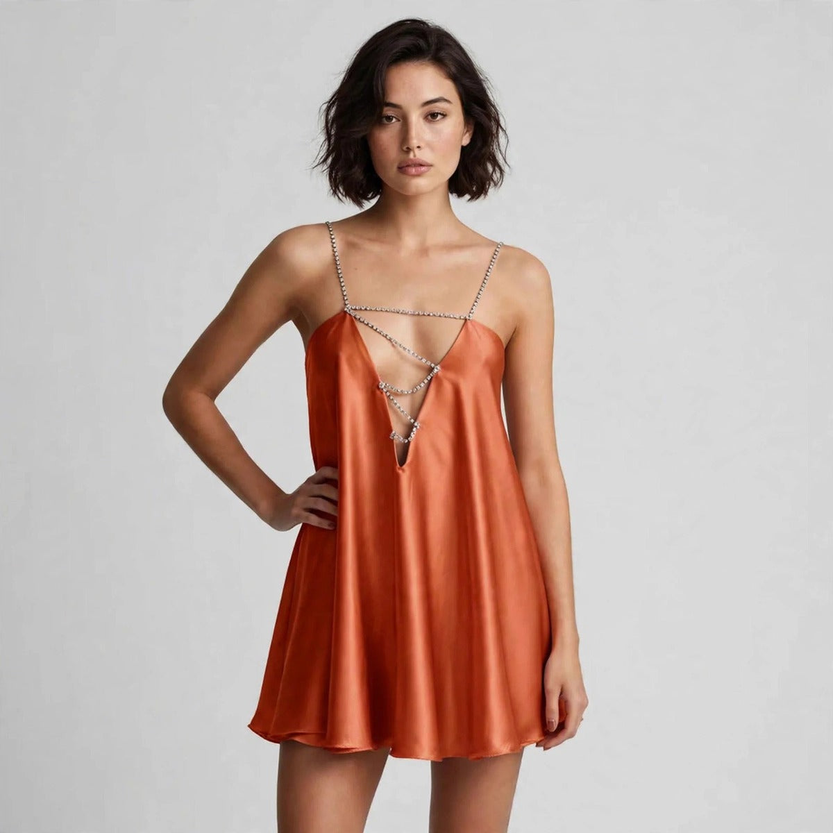 Woman posing with hand on hip in a burnt orange satin slip mini dress with rhinestone straps and chain lace-up chest cutout.