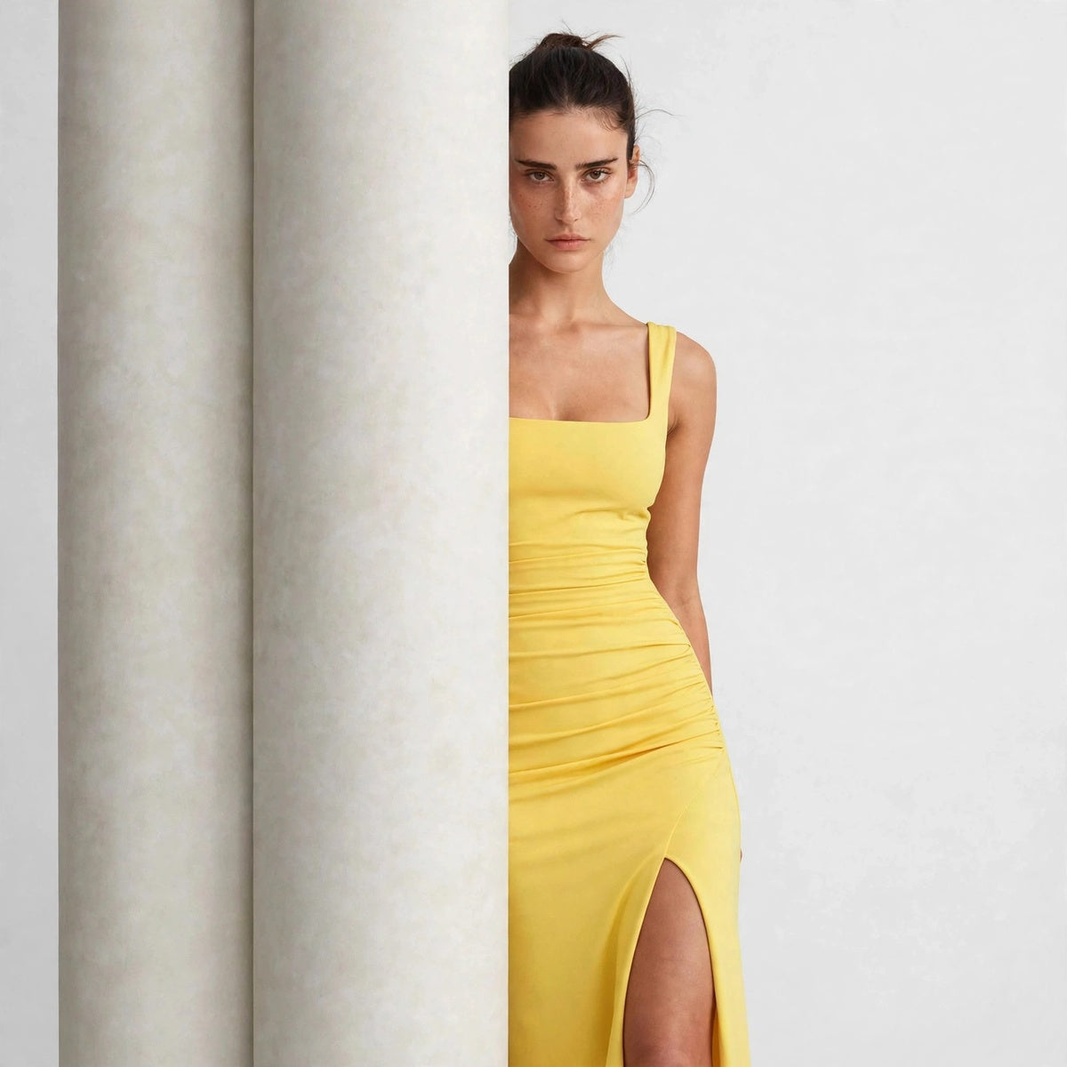 Model in a yellow ruched high-slit maxi dress peeking from behind a column, editorial view