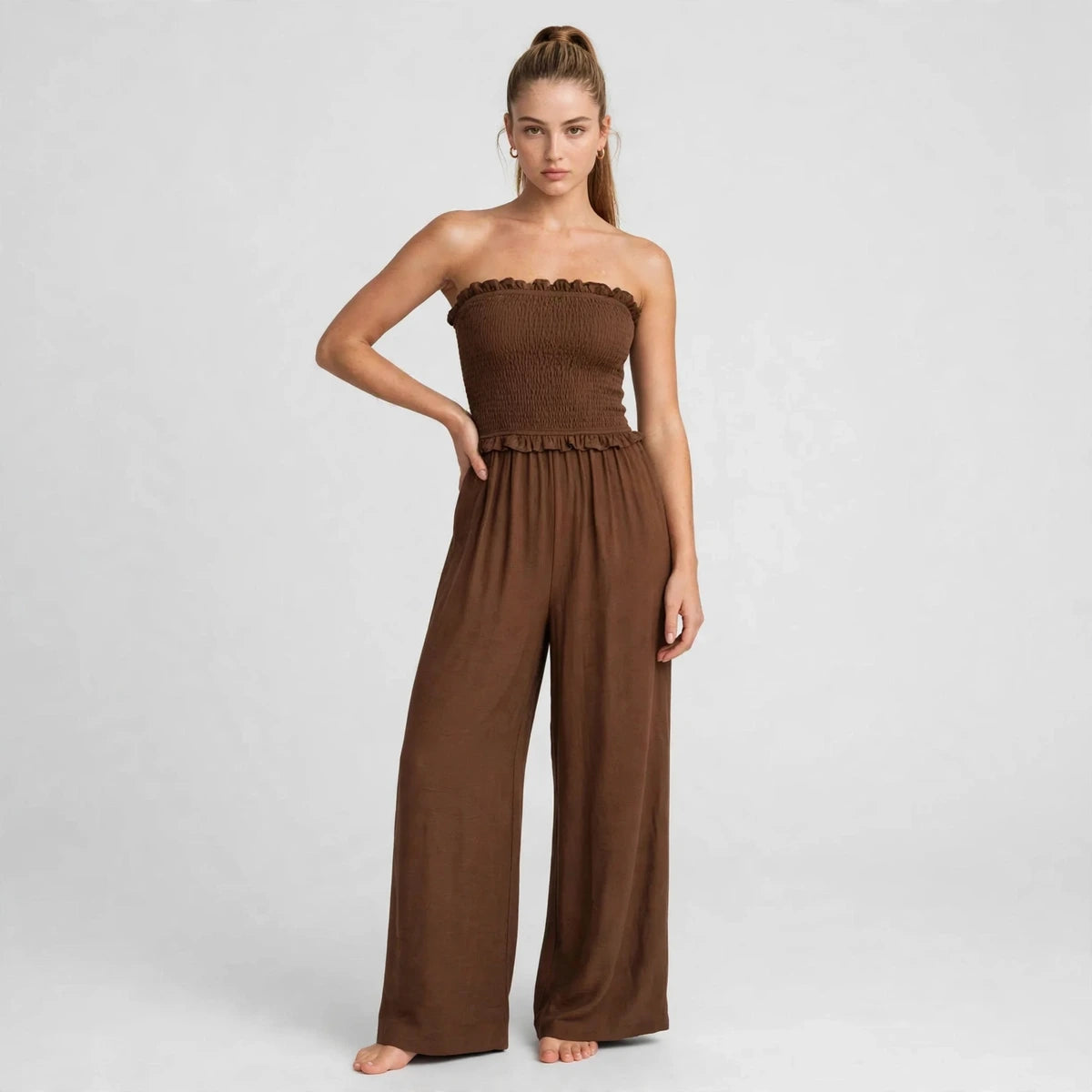 Woman wearing brown smocked strapless jumpsuit with ruffle trim and wide-leg pants, front view
